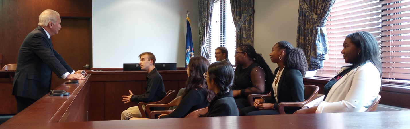 A group of young people sitting in a jury box while a man in a suit speaks to them.